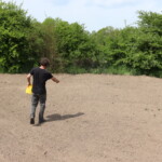 Ein Mann steht auf einem unbewachsenen Ackerboden und macht eine streuende Handbewegung, in der zweiten Hand ist ein Behälter