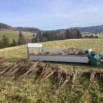 Blick über hügelige Wiesen- und Waldlandschaft, im Vordergrund liegende Bündel mit ca. 1m langen Heckenpflanzen