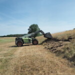 An einer Böschung, die an einer gemähten Wiese angrenzt, schaufelt ein Bagger die aus Gräsern bestehende Vegetation ab.