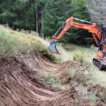 Ein Bagger steht auf einem Feld-/Waldweg und schaufelt an der linken Wegböschung die Vegetation bis auf den rohen Boden ab.