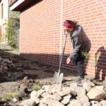 Frau mit Spaten auf Fläche mit Steinhaufen vor Hauswand