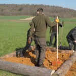Menschen mit Schaufeln stehen in einem von Holzstämmen umsäumten Sandarium