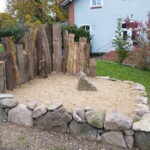 Ein etwa 2 x 2 Meter großer Sandkasten, vorne mit großen Steinen eingefasst und an der Rückwand mit aufrechten Baumstämmen eingerahmt