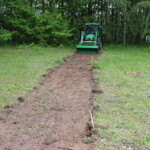 Ein Streifen mit bloßer Erde, ca. 1,20 Meter breit und ca. 10 Meter lang, auf einer Wiese, am Ende des Streifens ein grüner Traktor mit abgesenkter Schaufel in der Breite des Erdstreifens.