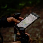 Zu sehen sind die Unterarme und Hände einer Person, die Wanderstöcke und ein Smartphone hält. Auf dem Display ist eine digitale Karte geöffnet. Der dunkle Hintergrund deutet auf einen schattigen Waldweg hin.