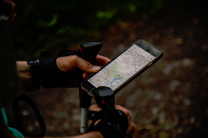 Zu sehen sind die Unterarme und Hände einer Person, die Wanderstöcke und ein Smartphone hält. Auf dem Display ist eine digitale Karte geöffnet. Der dunkle Hintergrund deutet auf einen schattigen Waldweg hin.
