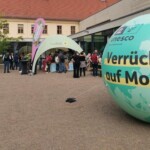 Im rechten Vordergrund des Bildes liegt eine etwa zwei Meter hohe Weltkugel, die mit dem UNESCO-Logo sowie der Aufschrift "Verrückt auf Morgen" verziert ist. Im Hintergrund sind mehrere Personen zu sehen, die an einem Stand miteinander ins Gespräch vertieft sind.
