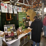 Ein Stand betreut von einer Mitarbeiterin der Verwaltungsstelle der Biosphärenverwaltung Berchtesgadener Land mit bunten Postkarten, Flyern, Wildkräuterstempel, Samentüten, selbstgemalten Postern mit bunten Blumen und vielen weiteren Infomaterialien. Davor mehrere interessierte Besucher*innen.