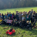 Gruppenfoto einer Schulklasse mit vier Betreuer*innen auf einer Wiese. Auf der Wiese liegen Gießkannen, Schnur, Draht und eine rote Plastebox – alles Dinge, die man für eine Pflanzaktion benötigt.
