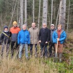 Sechs Männer und eine Frau stehen am Rande eines Nadelwaldes und lächeln in die Kamera für ein Gruppenfoto.