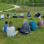 Eine Gruppe von Personen sitzt im Kreis auf einer grünen Wiese. Im Hintergrund erstrecken sich ein Weg und ein Teich.