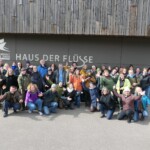 Eine Gruppe von Personen hat sich vor einem Gebäude mit der Aufschrift „Haus der Flüsse“ zu einem Gruppenbild aufgestellt und blickt in die Kamera, die Personen in der ersten Reihe befinden sich in der Hocke.