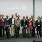 14 erwachsene Personen stehen für ein Gruppenbild zusammen, im Hintergrund an der Wand das Logo „Nationale Naturlandschaften“, rechts und links Aufsteller mit dem Schriftzug „Naturschutz im Urlaub“.