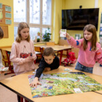 Vier Kinder spielen an einem Tisch mit einem bunten Wimmelbild sowie verschiedenen Karten.