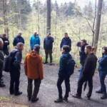 In einem Wald stehen Menschen in einem Kreis zusammen und hören einer Person aus ihrer Mitte zu.
