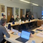 Rings um in einem Viereck aufgestellte Tische sitzen Menschen und reden miteinander. Im Hintergrund ist über einen Monitor eine Person zugeschaltet.
