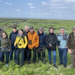 Auf einer Salzwiese im Wattenmeer stehen Menschen zu einem Gruppenfoto zusammen. Am Horizont ist als Streifen die Nordsee zu sehen.