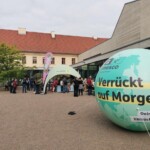 Im Vordergrund rechts befindet sich ein großer, grüner Ballon in Form eines Globus, auf dem "UNESCO Biosphärenreservat" und "Verrückt auf Morgen" steht. In der Bildmitte ist ein Informationsstand unter einem Pavillon, an dem sich mehrere Menschen aufhalten. Dahinter ist ein Gebäude zu sehen.