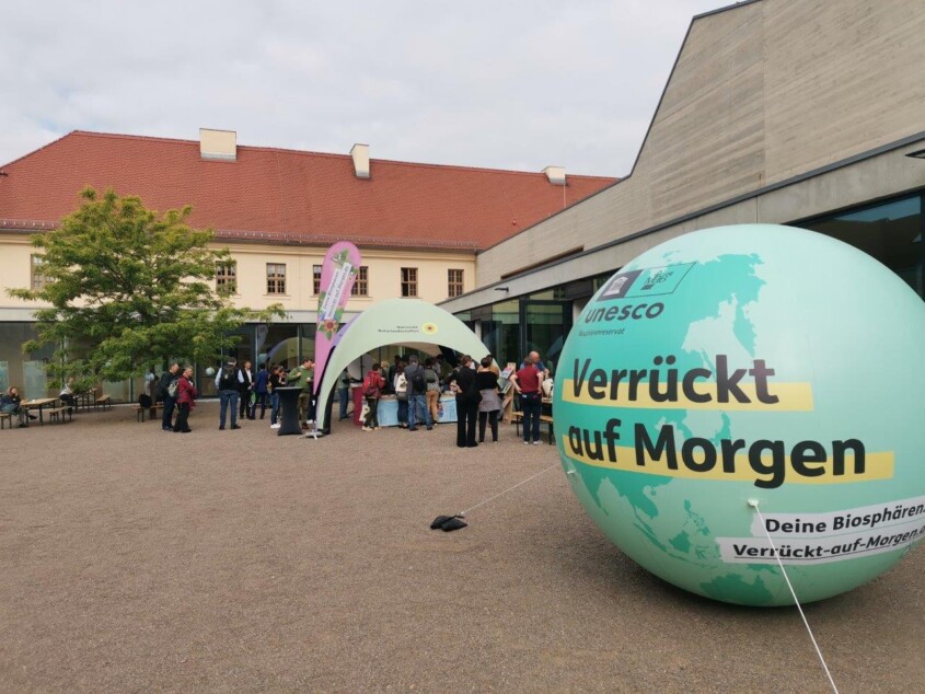 Im Vordergrund rechts befindet sich ein großer, grüner Ballon in Form eines Globus, auf dem "UNESCO Biosphärenreservat" und "Verrückt auf Morgen" steht. In der Bildmitte ist ein Informationsstand unter einem Pavillon, an dem sich mehrere Menschen aufhalten. Dahinter ist ein Gebäude zu sehen.