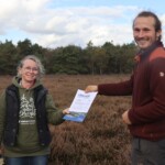 Ein Mann überreicht einer Frau eine Urkunde, im Hintergrund Heide und Wald.