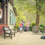 : Eine Frau winkt einer Familie mit zwei Kindern zu, die mit Gepäck auf dem Hof eines Gasthauses ankommen. Im Vordergrund ruht ein schwarzer Hund entspannt auf dem Boden.