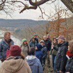 14 Erwachsene stehen in einer Gruppe um einen Redner zusammen, im Hintergrund bewaldete Berghänge und ein großes Gewässer.