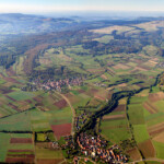 Landschaftsaufnahme aus der Vogelpersketive mit Siedlungen, Waldstücken und grünen und braunen Flurstücken im Wechsel.