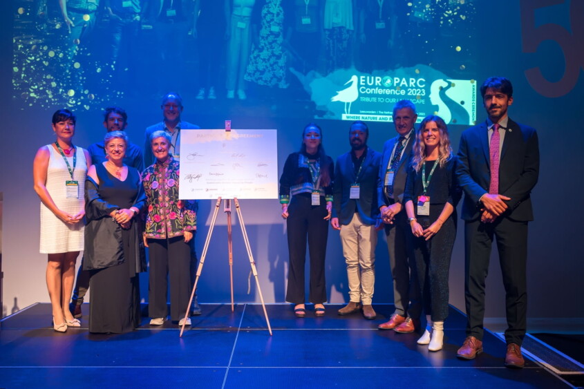 Auf dem Foto sind zehn Frauen und Männer zu sehen – Vertreter*innen der acht Ländersektionen, die Sektionssprecherin sowie der Präsident der EUROPARC Federation. Sie stehen auf einer Bühne, angestrahlt von Scheinwerfern, zwischen ihnen die neue Partnerschaftsvereinbarung, symbolhaft auf einer Tafel abgebildet, auf der alle Sektionsvertreter*innen vorab unterschrieben haben.