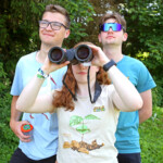 Drei junge Menschen blicken gemeinsam auf einen Punkt am Horizont hinter der Kamera. Das Mädchen schaut dabei durch ein Fernglas.