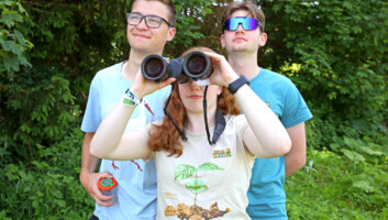Drei junge Menschen blicken gemeinsam auf einen Punkt am Horizont hinter der Kamera. Das Mädchen schaut dabei durch ein Fernglas.