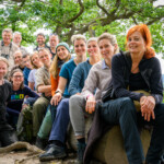 Eine Gruppe von Menschen sitzt in einem Wald hintereinander auf einem liegenden Baumstamm, einige stehen und hocken drumherum. Alle lächeln in die Kamera. Eine Gruppe von Menschen sitzt in einem Wald hintereinander auf einem liegenden Baumstamm, einige stehen und hocken drumherum. Alle lächeln in die Kamera.