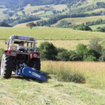 Ein Trecker mit Balkenmäher bei der Arbeit in einer hügeligen und grünen Landschaft.
