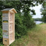 Am Rande einer Uferböschung ist eine dreiteilige Insektennisthilfe aus Holz aufgestellt. Seitig des Holzkastens hängt eine Erklärtafel mit Bildern.