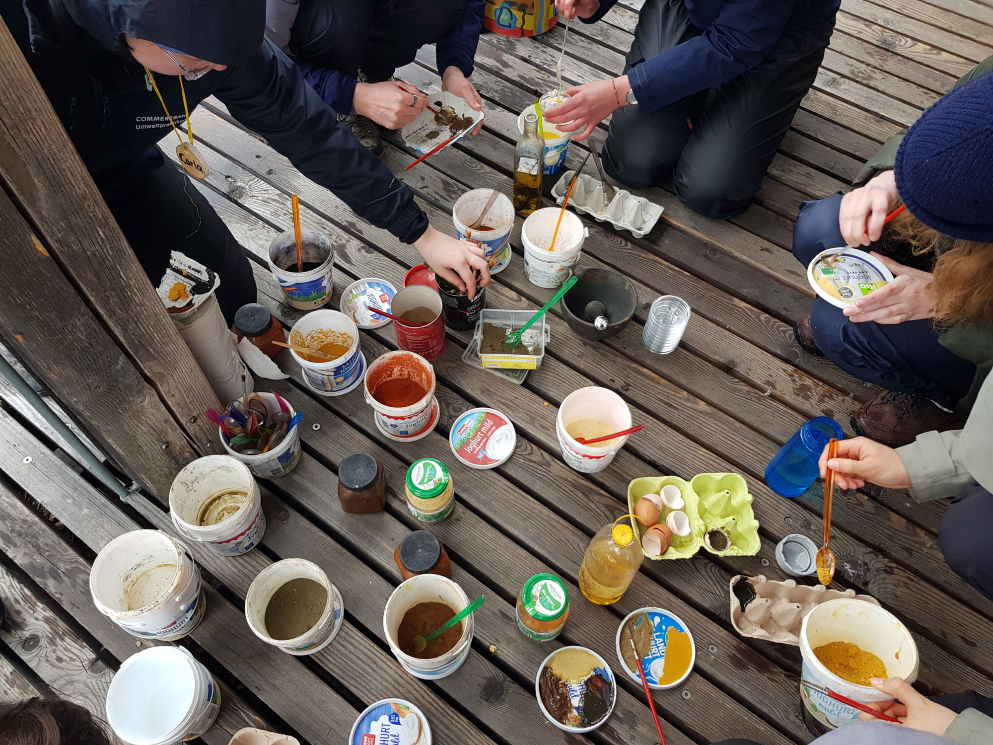 auf dem Boden einer Holzterasse stehen Flaschen, Eierkartons und viele recycelte Becher mit bunten Farben, angemischt aus Pigmenten und Materialien aus der Natur