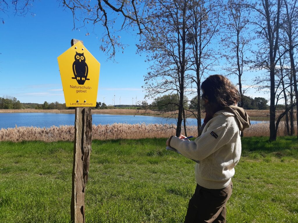 Ein Person rechts im macht sich Notizen auf einem Block. Link im Bild ist ein Natuschutzgebiet-Schild zu sehen, im Hintergrund ein Gewässer und Schilf.
