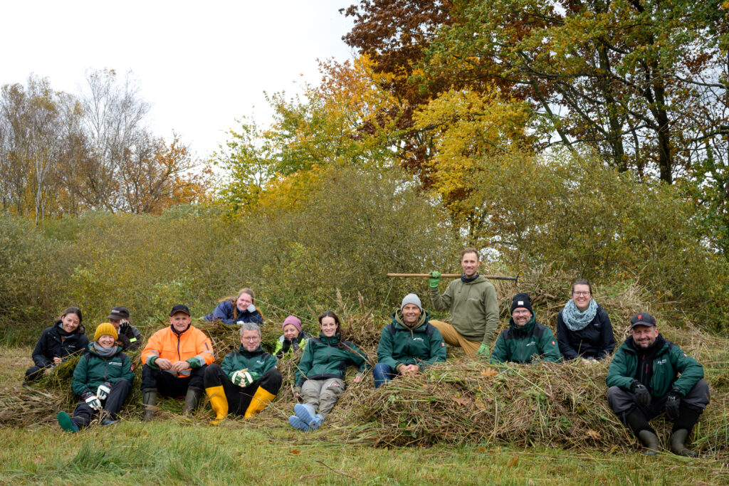 Dreizehn Erwachsene in Arbeitskleidung und mit Harke auf einem Berg von Mähgut