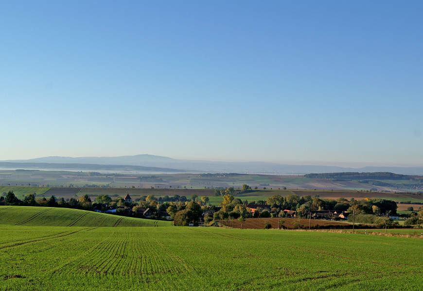 Eine landwirtschaftlich genutzte Hügelfläche leitet den Blick in ein Tal mit Bäumen und Dörfern.