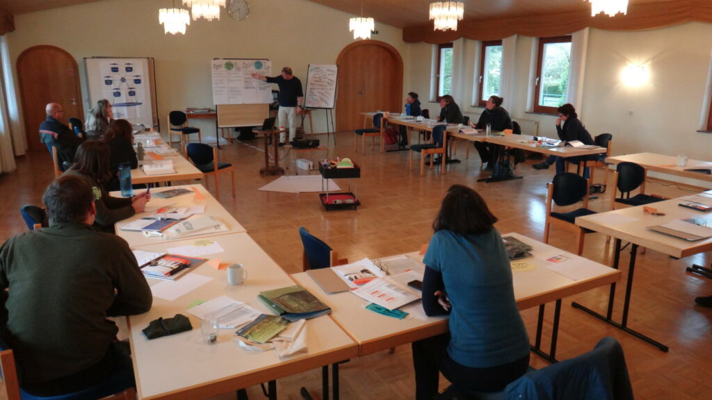 Seminarraum mit etwa 10 Personen an Tischen in U-Form und zeigende Person vorne an Pinnwand