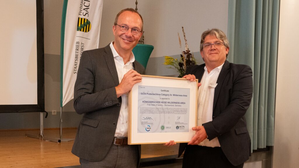 Der rechts stehende Mann, der deutsche IUCN-Vertreter Dr. Eick von Ruschkowski, überreicht dem links stehenden Mann, Sachsens Umweltminister Wolfram Günther, die Urkunde über die Auszeichnung der Königsbrücker Heide als IUCN-Wildnisgebiet
