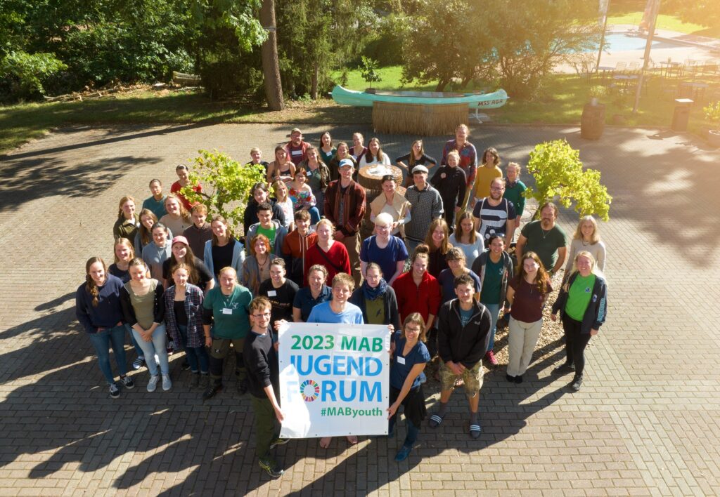 Teilnehmende des MAB Jugendforums gruppieren sich um einen Brunnen und halten ein Banner mit dem Logo MAB Jugendforum 2023 hoch.