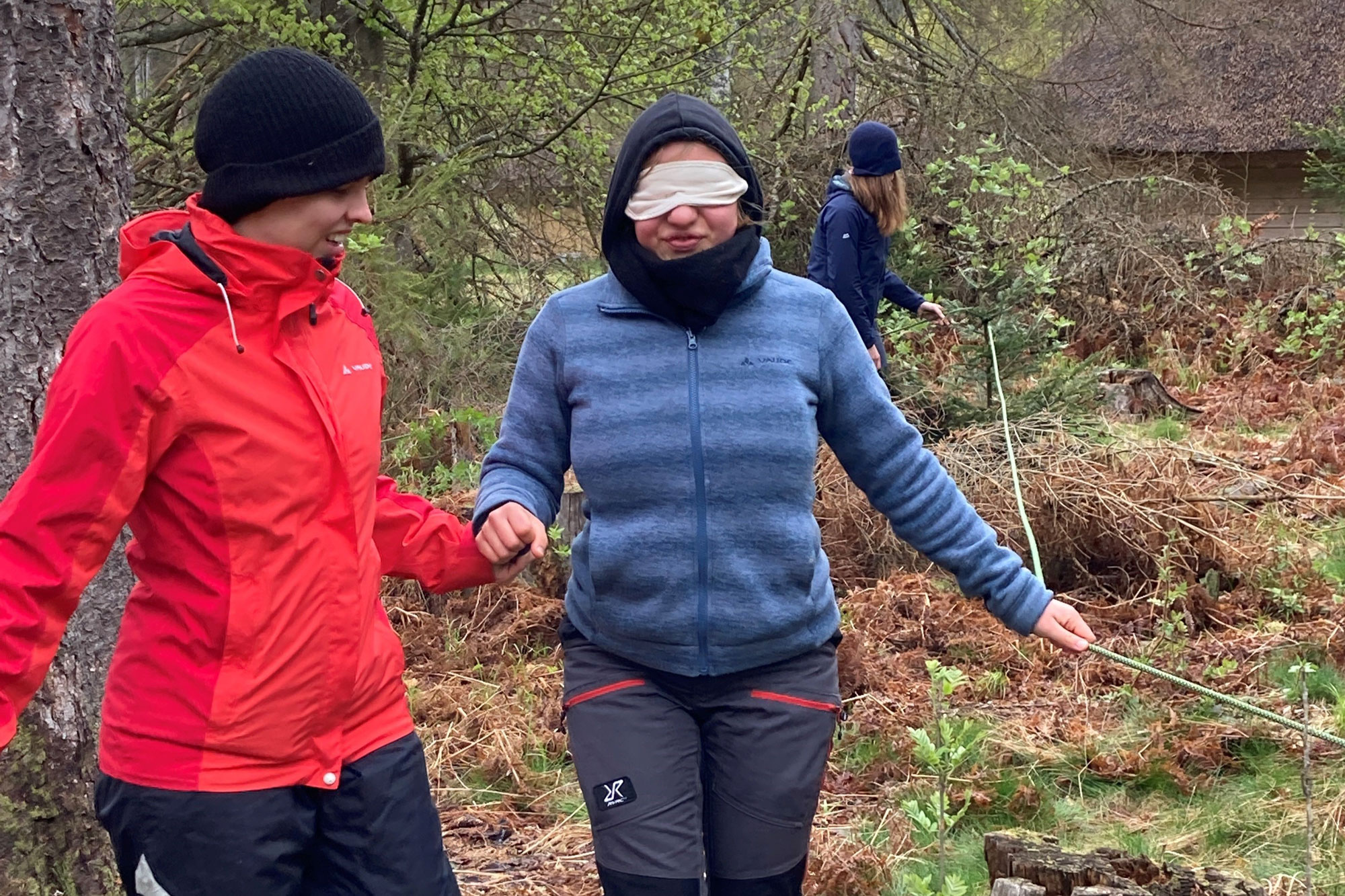 eine Person in roter Jacke und schwarzer Mütze führt eine Person in blauer Jacke und verbundenen Augen einen Weg im Wald entlang