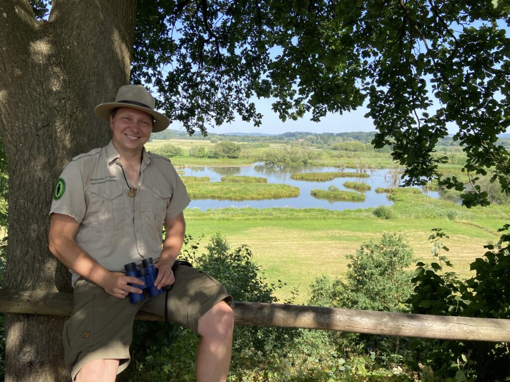 Ein Ranger sitzt lächelnd auf einem Holzgeländer, im Hintergrund eine Wasser-Wiesen-Landschaft