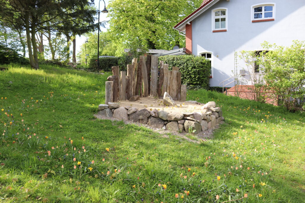 Sandiger Bereich umrandet mit Steinen und Totholz auf einer Wiese mit Frühblühern