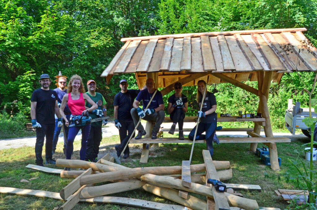 Gruppe aus 8 Personen vor und neben einem hölzernen Picknickplatz mit Bedachung, davor Bauholz auf dem Boden liegend