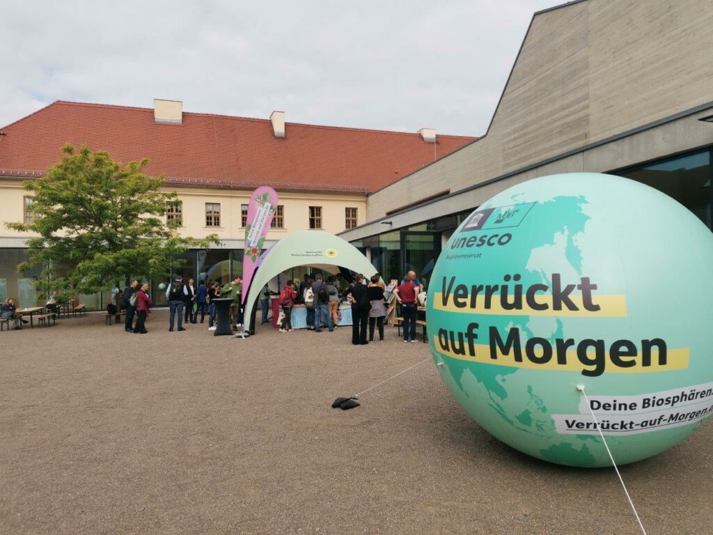 Im Vordergrund rechts befindet sich ein großer, grüner Ballon in Form eines Globus, auf dem "UNESCO Biosphärenreservat" und "Verrückt auf Morgen" steht. In der Bildmitte ist ein Informationsstand unter einem Pavillon, an dem sich mehrere Menschen aufhalten. Dahinter ist ein Gebäude zu sehen.